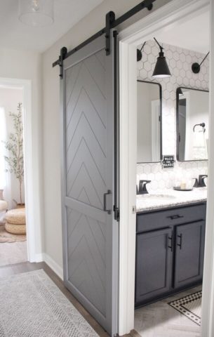 Grey herringbone barn door with a barn door lock leading into a modern white bathroom.