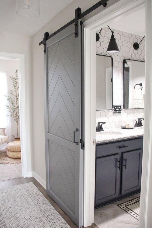Grey herringbone barn door with a barn door lock leading into a modern white bathroom.