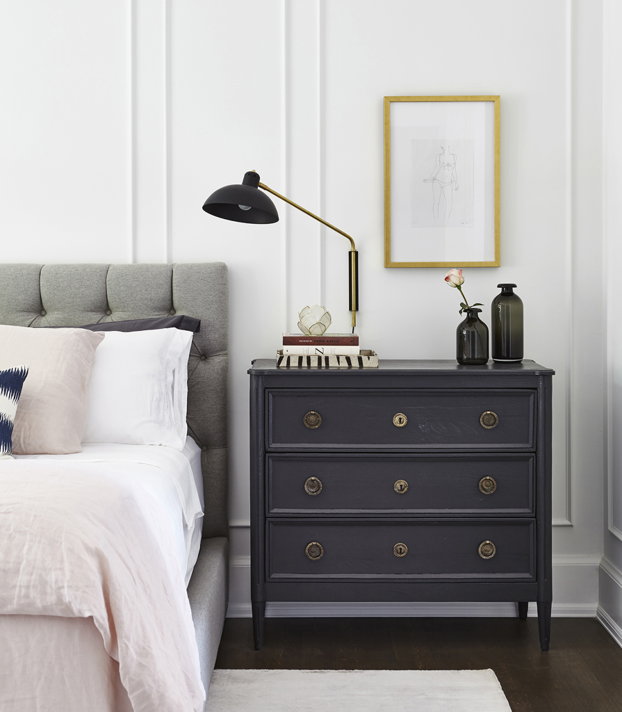 Black three drawer side table next to grey bed in a modern bedroom.
