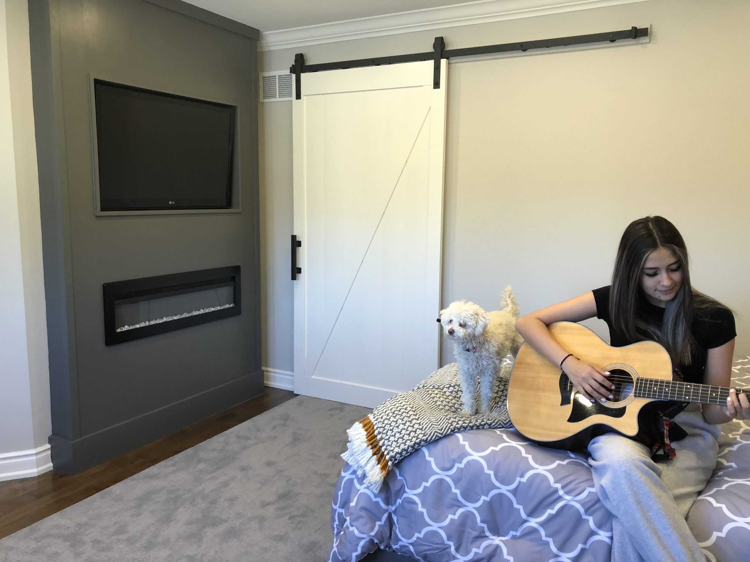 White Barn Door behind teen girl sitting with a small white dog playing guitar on her bed.